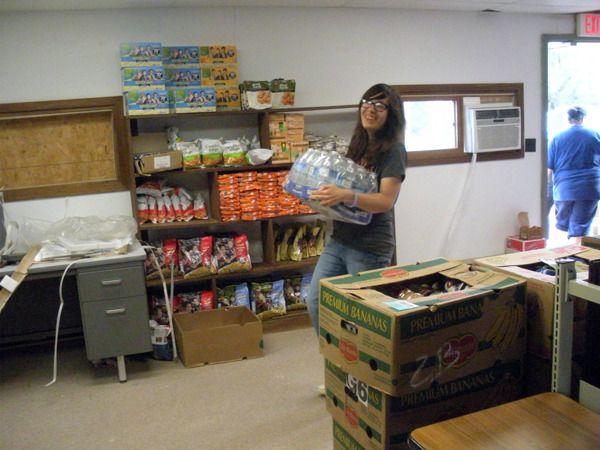 Restocking Okiciyap Food Pantry, Smiling woman carrying a case of bottled water to stack on the shelves in the Okiciyap Food Pantry. One of the new window a/c units is in the background.